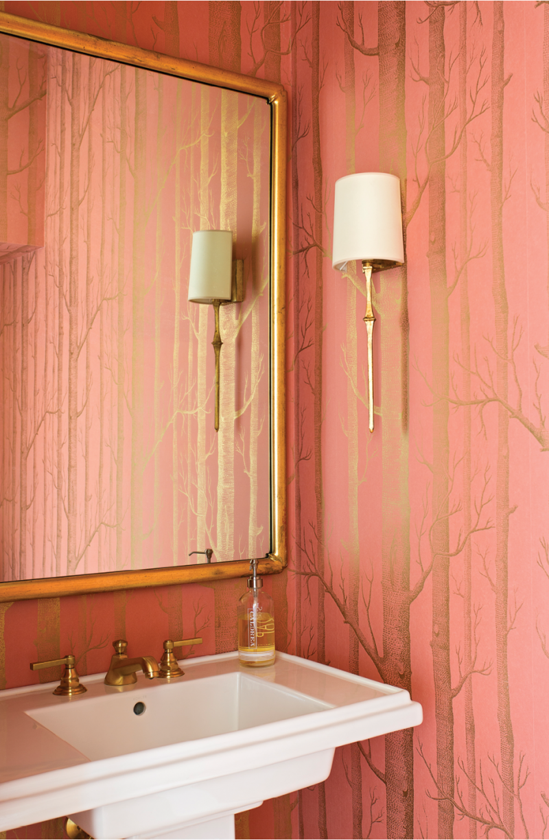 The powder room dazzles in radiant gold-flecked Cole &amp; Son wallpaper, accented by a mirror found at John Pope Antiques and delicate sconces from Circa Lighting.