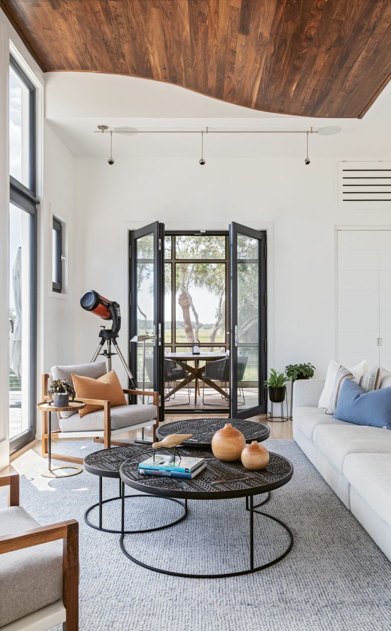 Tasked with creating the smallest house plausible, architect Myles Trudell opted to put the dining room outside, in a screened porch. While the surroundings take center stage in the adjacent living room, pops of visual interest are provided by natural textures, such as a spectacular walnut ceiling feature and three Tabwa nesting coffee tables from Ethnicraft, whose blackened teak tops are carved in geometric reliefs.